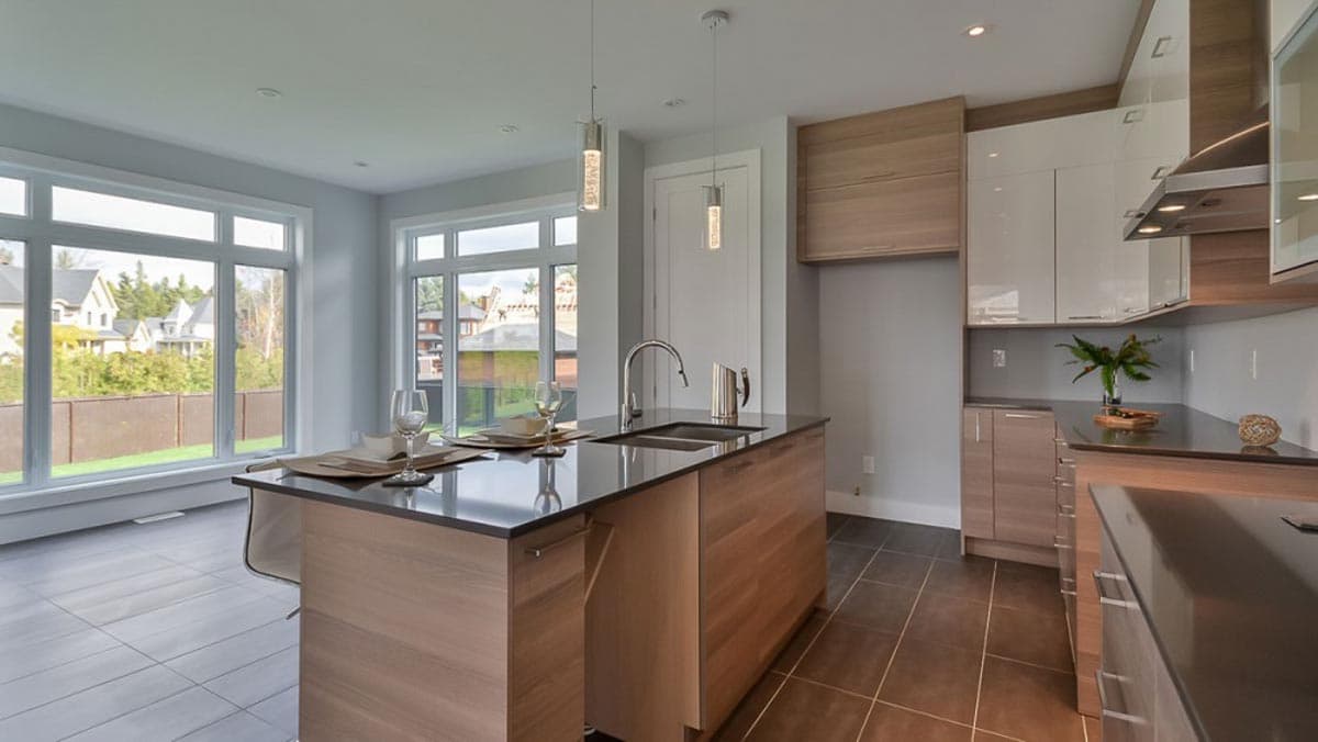 Modern kitchen with island, double sink, dark countertops, and white and wood cabinetry. Large windows overlook backyards.