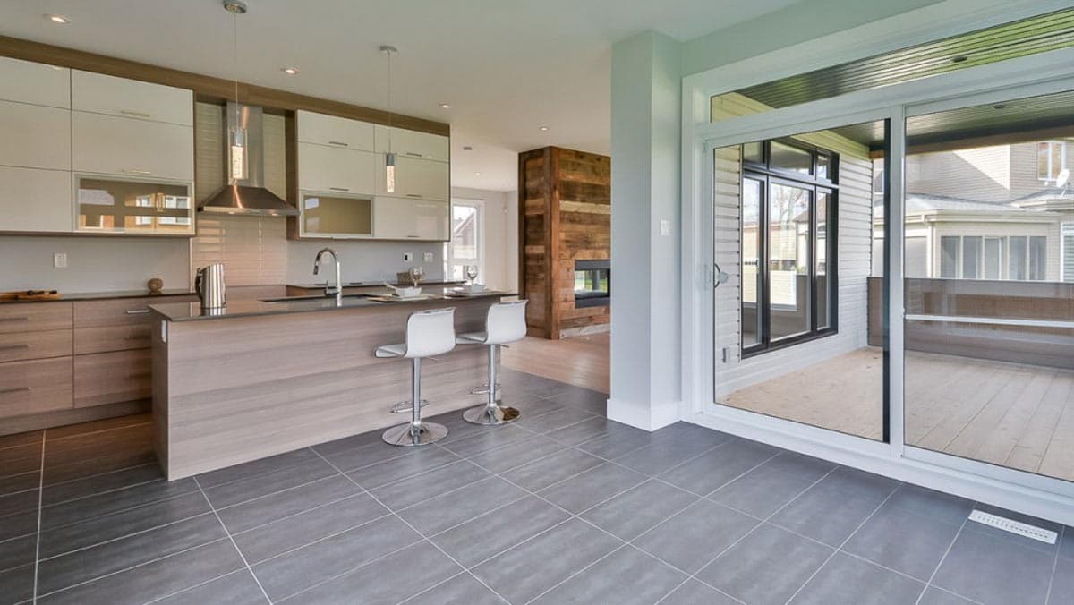 Modern kitchen with island, stainless steel appliances, and adjacent great room with wood-paneled fireplace. Sliding glass doors open to deck.