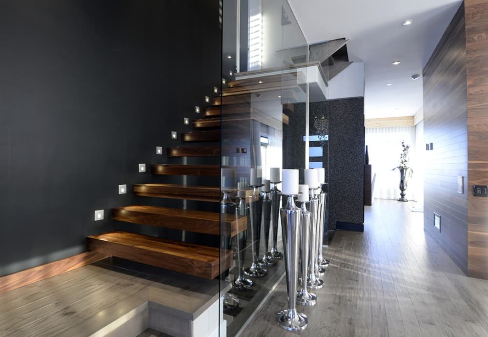 Modern staircase with floating wood treads, glass railing, and wall-mounted lights. Hallway with wood paneling.