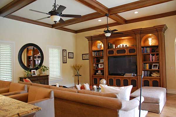 Living room with coffered wood beam ceiling, ceiling fans, built-in entertainment center, and large windows with shutters.