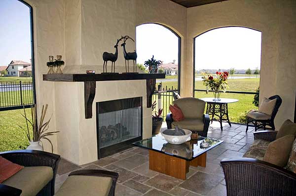 Covered outdoor living space with fireplace, seating, and arched openings to a landscaped yard.