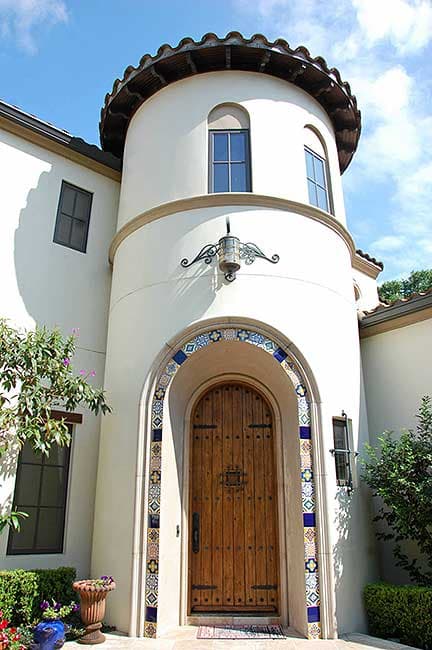 House plan exterior with a stucco finish, arched wooden door with decorative tile surround, and a cylindrical tower with a tile roof.