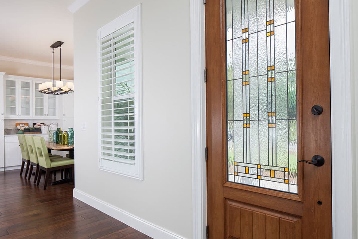 Interior view of a house entryway with a stained-glass door and plantation shutters next to a dining area with cabinets and a chandelier.