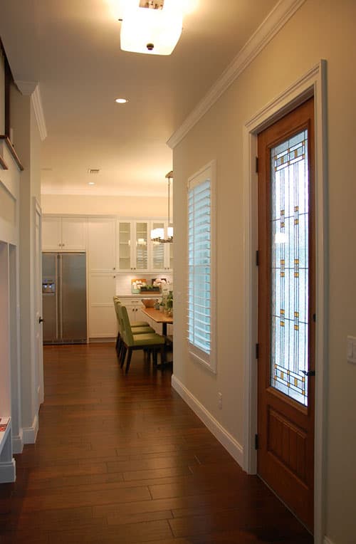 Hallway with stained glass front door, leading to kitchen with white cabinets, stainless steel refrigerator, and dining area with green chairs.