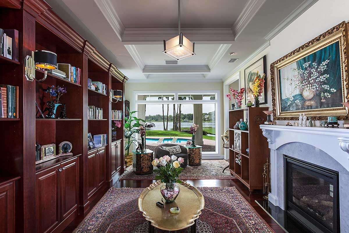 Interior view of a room with floor-to-ceiling bookshelves, a marble fireplace, and a large window overlooking a pool and lake.