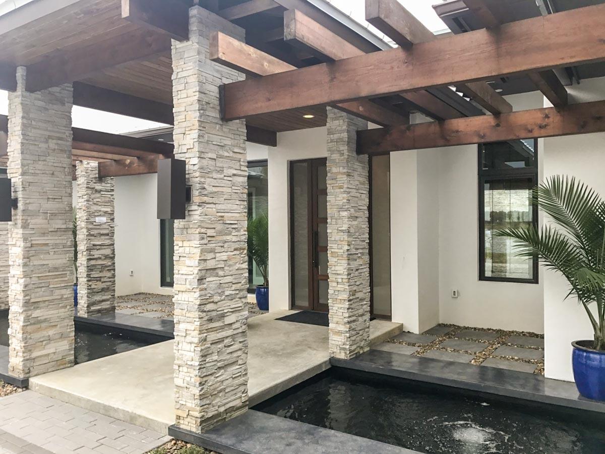 Modern house exterior with stacked stone pillars, wood beam pergola, water features, and large windows.