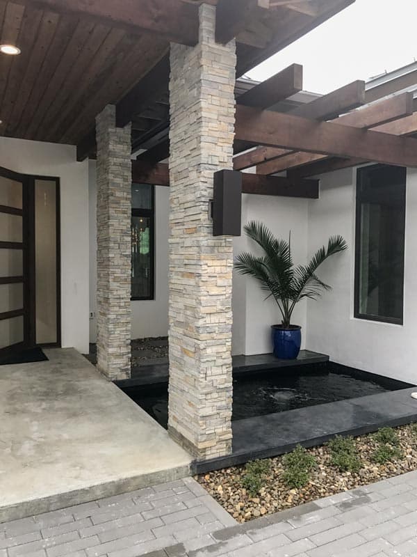 Modern house exterior with stacked stone pillars, wood pergola, water feature, and large windows.