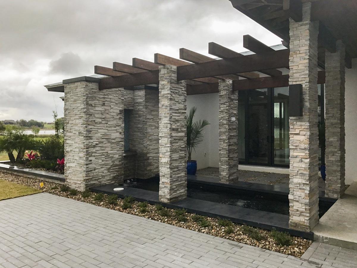 Modern exterior house plan featuring stone pillars supporting a wood-slat pergola over a water feature.