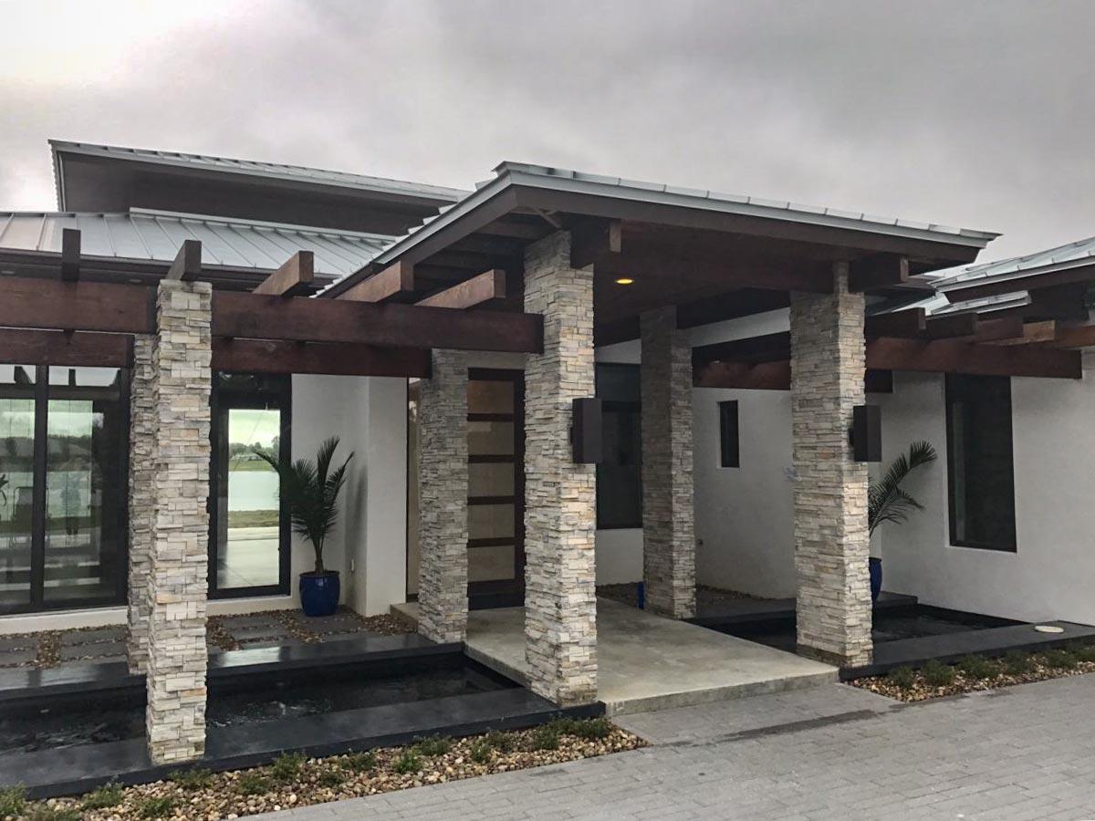 Modern house exterior with stone pillars, wood beams, pitched metal roof, and water features.