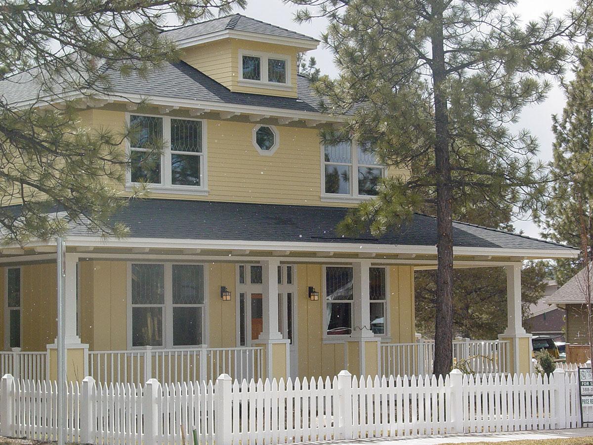 Two-story house plan exterior with a wraparound porch, gables, dormer windows, and a white picket fence.