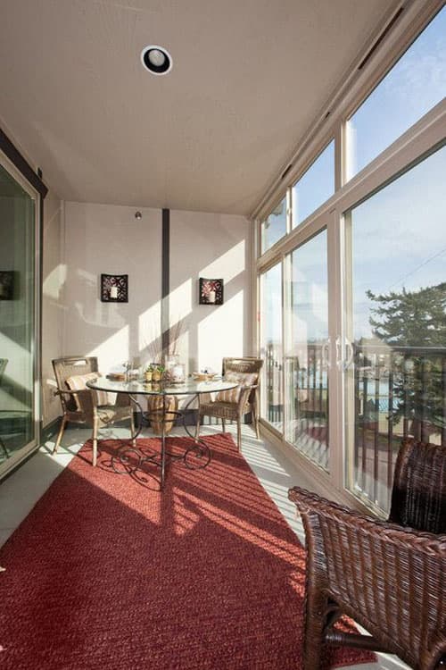 Sunlit enclosed patio with glass-top dining table, wicker chairs, and floor-to-ceiling windows overlooking a balcony.