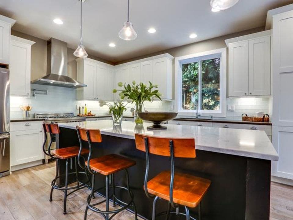 Kitchen with dark island, white cabinets, stainless steel range hood, and marble countertops. Seating for four at the island.