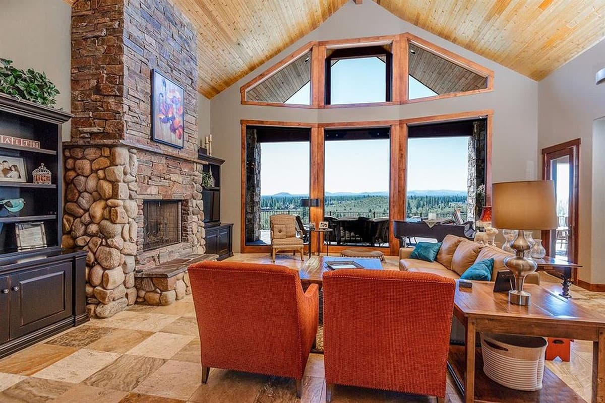 Great room interior with stone fireplace, vaulted wood ceiling, and large windows overlooking a scenic landscape.