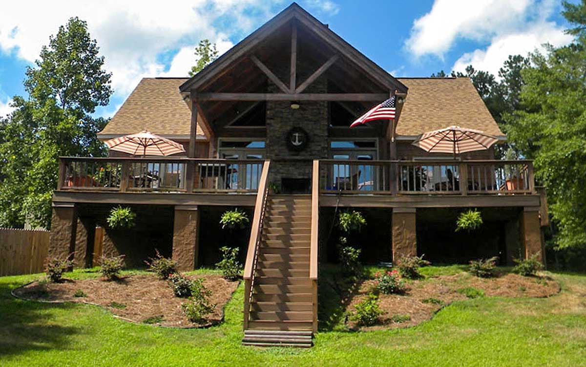 House plan exterior featuring a two-story design with a large wraparound deck, prominent gabled roof, and central stone chimney.