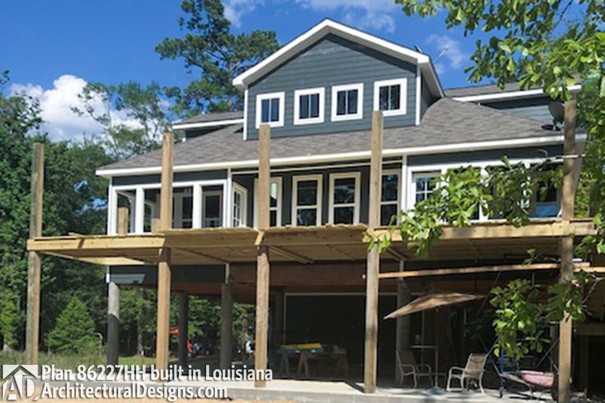 House plan exterior: Two-story home with gables, multiple dormers, and extensive wooden decking supported by posts.