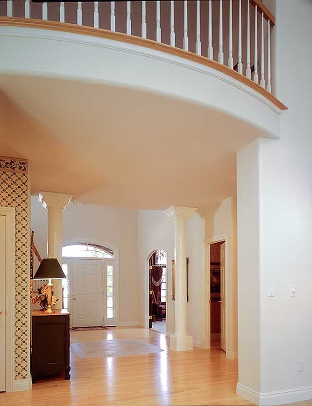 Interior view of a house entryway with a curved balcony, columns, arched doorway, and light wood floors.