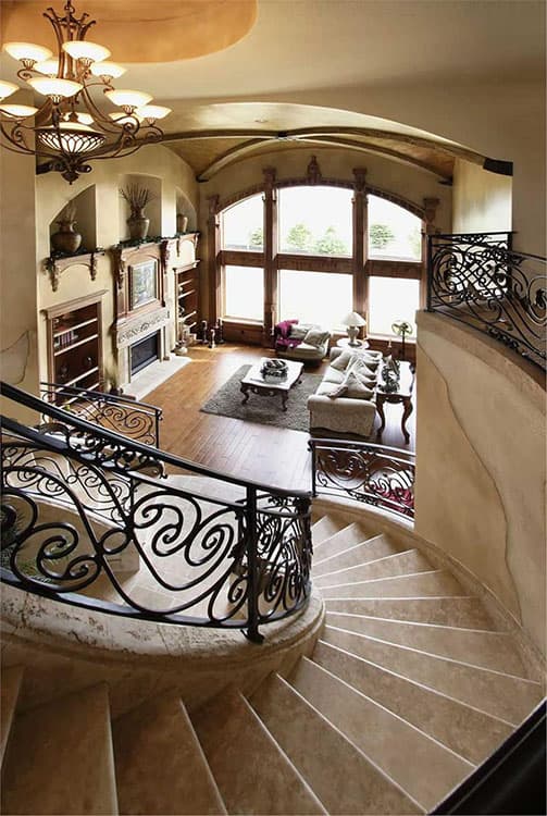 Interior view showcasing a curved staircase with wrought iron railing, leading to a living room with a fireplace and large arched windows.