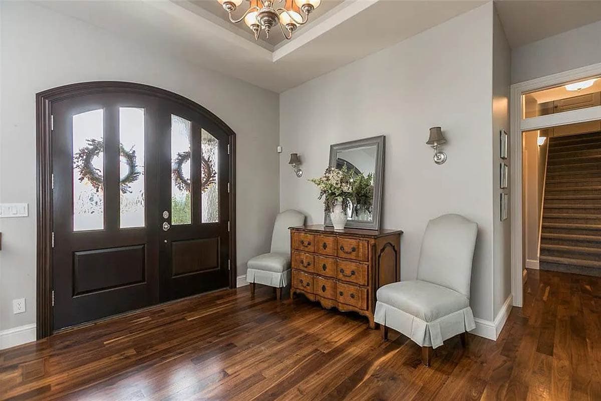 Interior view of entryway with double arched doors, dresser, two chairs, and staircase.