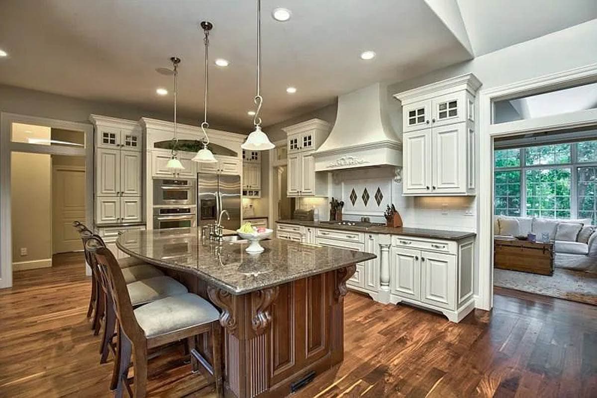 Interior view of a kitchen with a large island, stainless steel appliances, white cabinetry, and a pass-through to a living room.