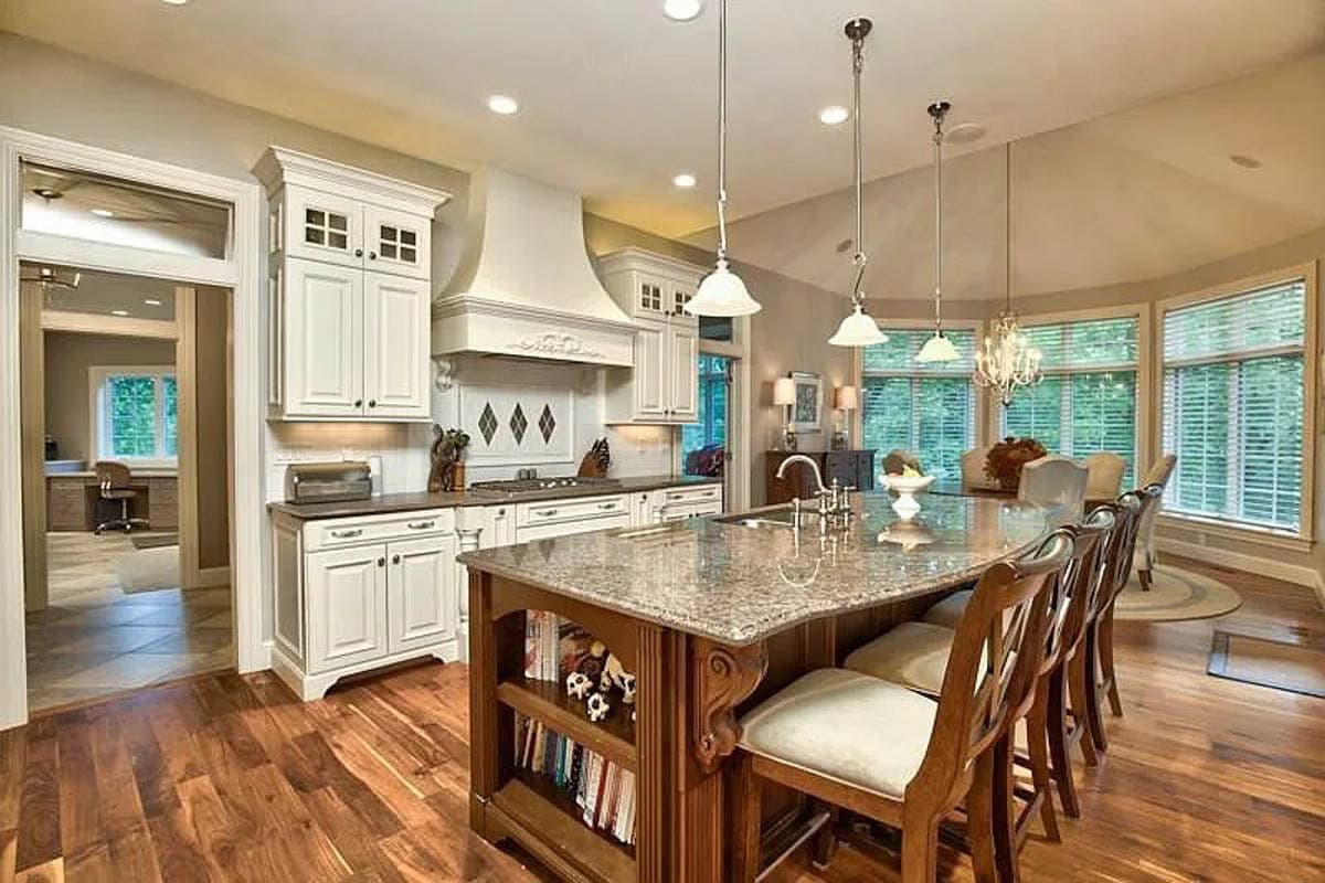 Kitchen with large island, granite countertops, white cabinetry, range hood, and dining area with bay windows.