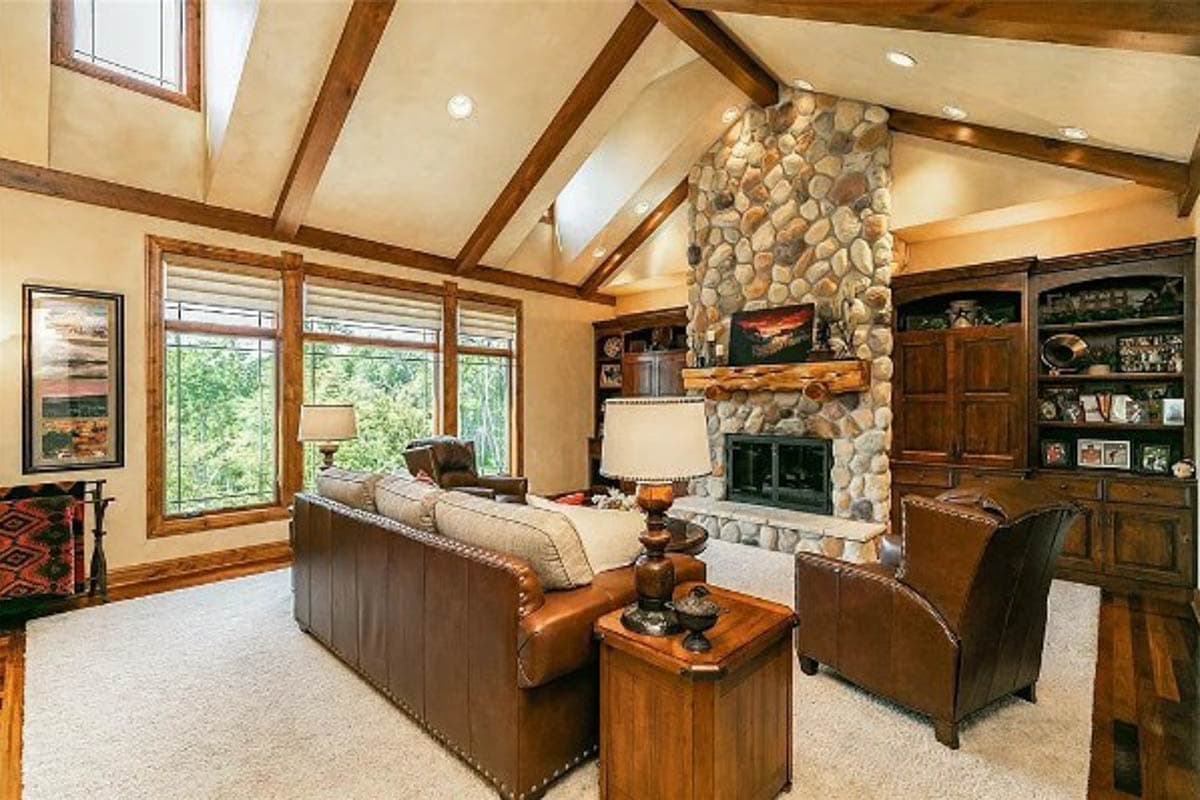 Living room with vaulted beamed ceiling, stone fireplace, built-in shelving, and large windows.