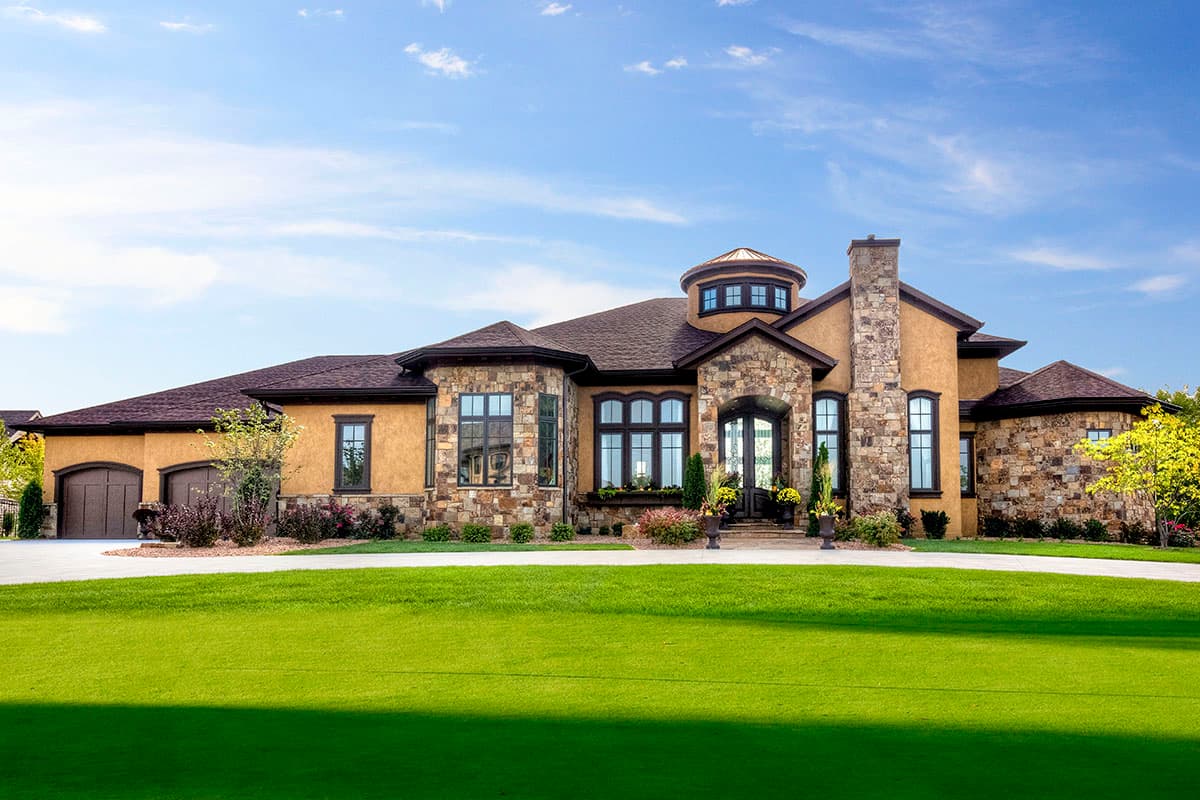 House plan exterior. Two-story with stone facade, turret, and a prominent chimney. Features include a three-car garage and gabled rooflines.