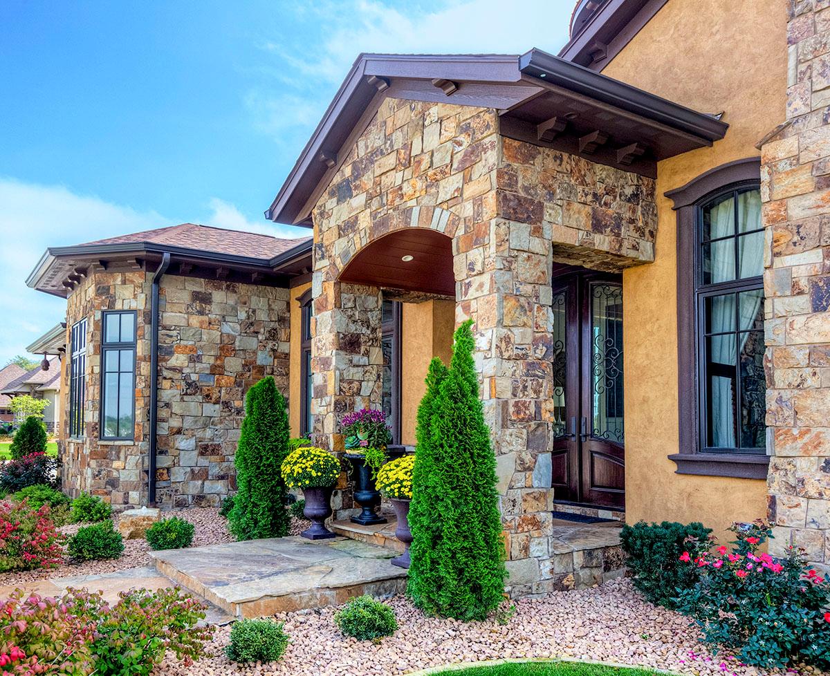 House plan exterior featuring stone facade, arched entry, multiple gables, and dark-framed windows.