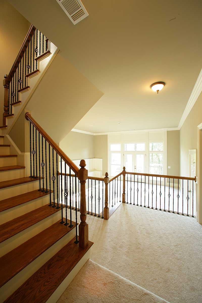 Interior view of staircase with wood treads and black metal balusters, leading to a carpeted landing with large windows.