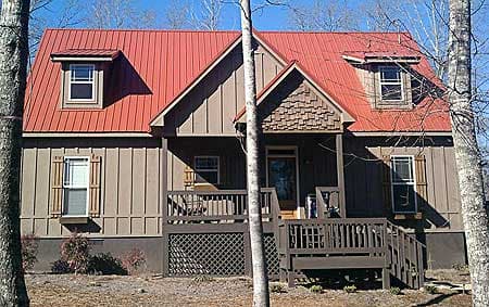 House plan exterior: Two-story home with red metal gable roof, dormer windows, wood siding, and a covered front porch with railing.