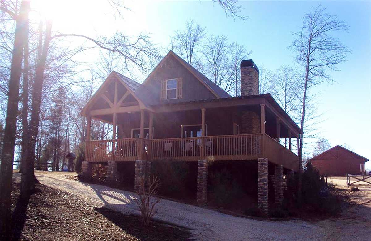 House plan exterior: two-story cabin with wraparound porch, exposed beams, metal roof, and stone chimney.