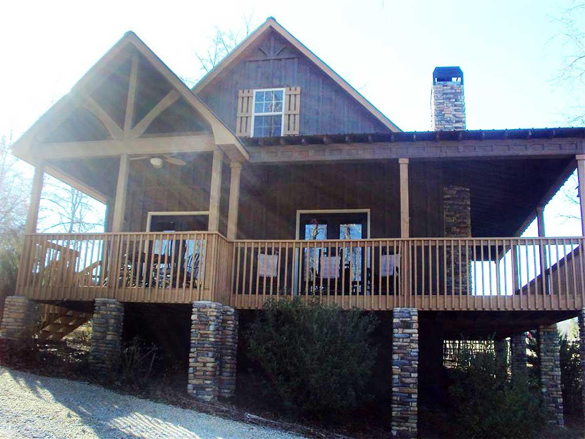 House plan exterior: Two-story home with prominent gables, stone chimney, and wraparound deck with wooden railings and stone piers.