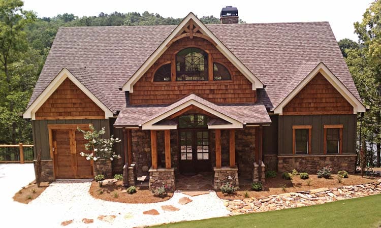 House plan exterior: Rustic style with stone and wood accents, covered front porch with timber posts, dormer with arched window, and attached garage.