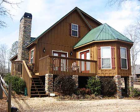 House plan exterior: Two-story wood-sided home with large stone chimney, wraparound deck, and multi-sided green metal roof.