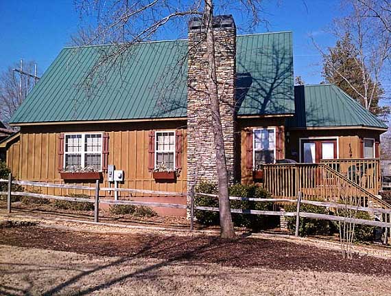 House plan exterior: Wood siding, green metal roof, stone chimney, wooden deck with stairs, and window boxes.