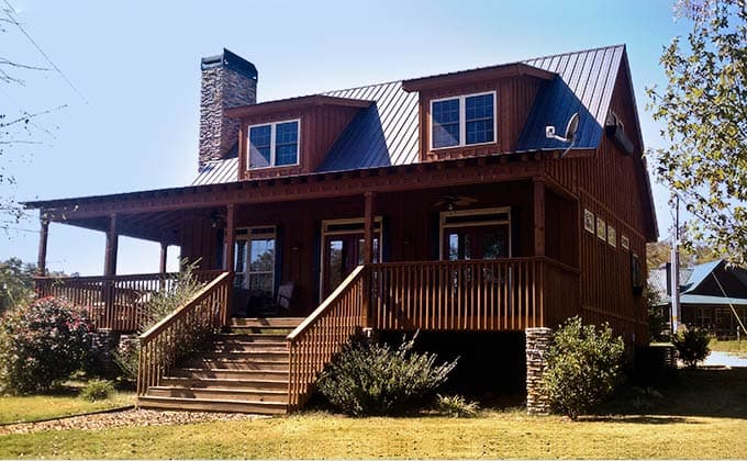 House plan exterior with gambrel roof, prominent stone chimney, and wraparound covered porch with deck.