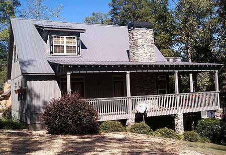 House plan exterior: Two-story cabin with metal roof, stone chimney, and wraparound porch with white railings.