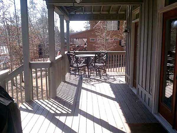 House plan exterior: Covered deck with dining set, wooden railing, and adjacent house structure visible.