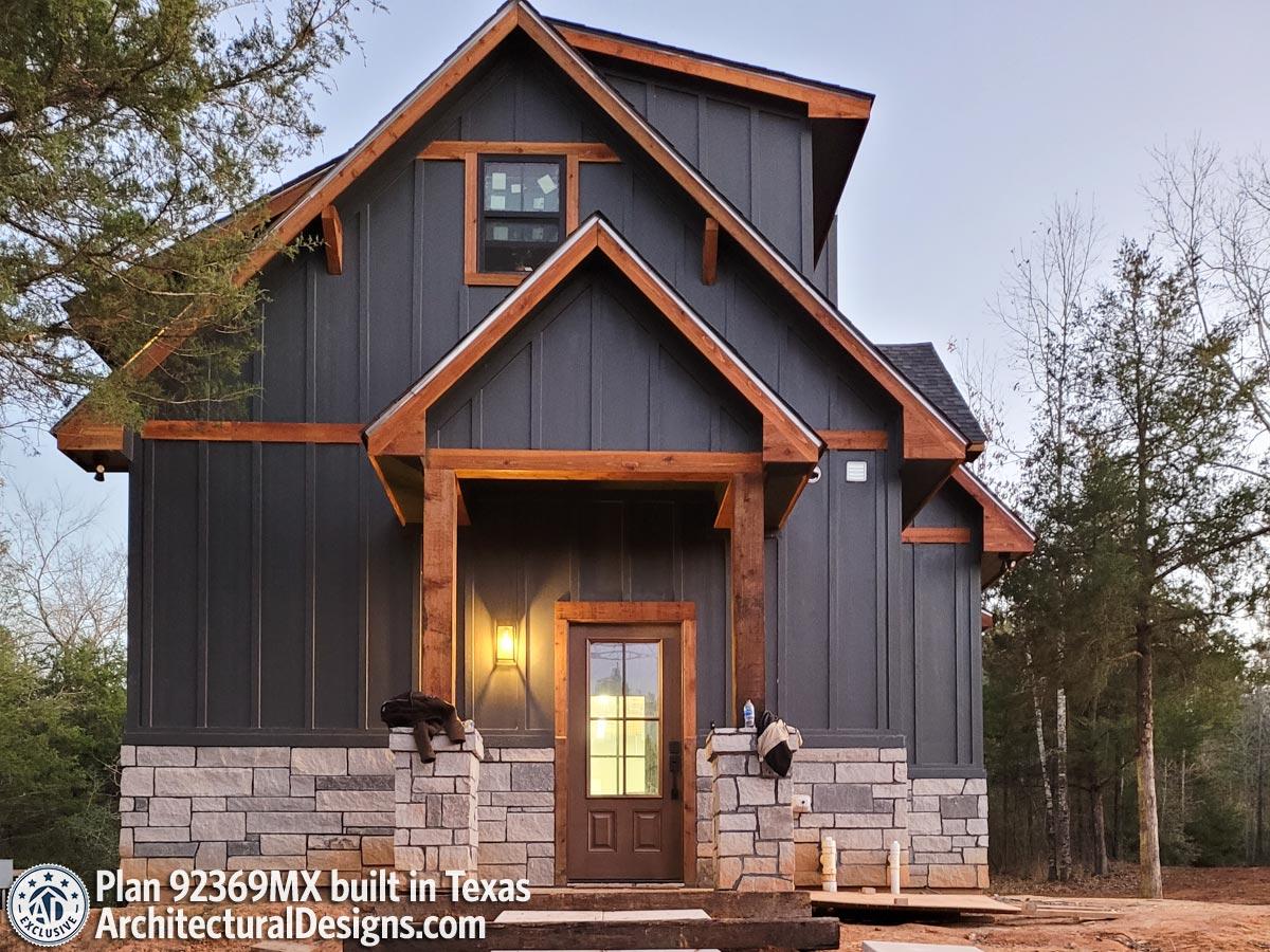 House plan exterior with dark board and batten siding, stone foundation, and exposed timber beams on entrance.
