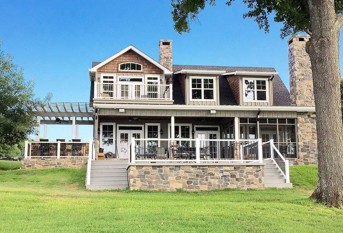 House plan exterior: two-story home with stone base, gabled dormers, upper balcony, and wraparound porch.