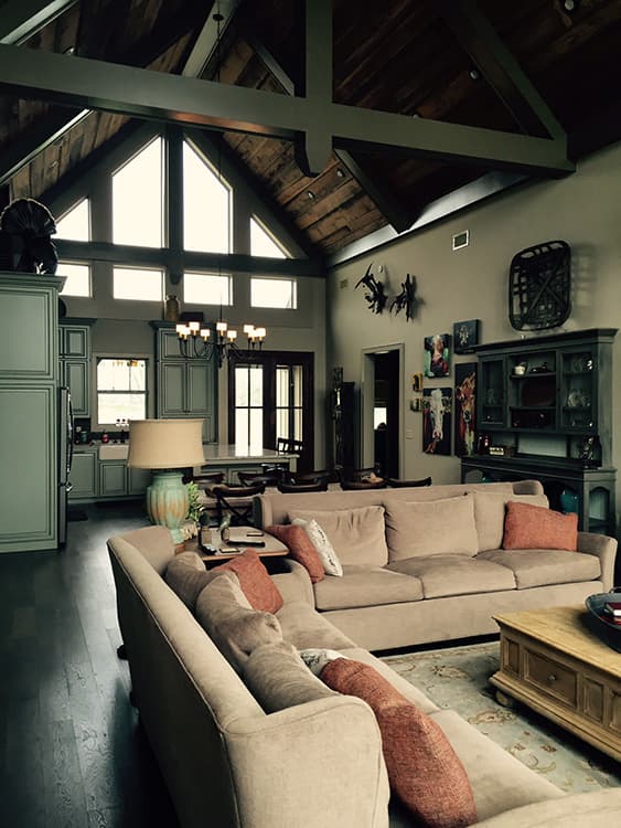 Living room with vaulted ceiling, exposed beams, large windows, sectional sofa, and kitchen area.