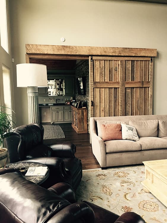 Living room with leather chairs, sofa, large rustic barn door, and view into a kitchen area with bar.