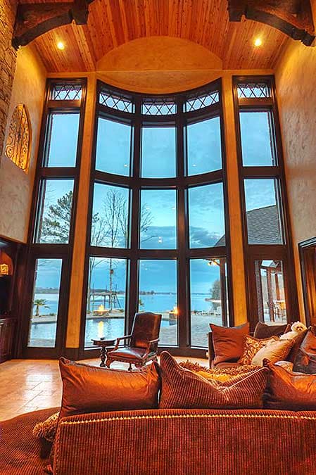 Interior great room with floor-to-ceiling arched windows overlooking a body of water. Timber ceiling beams and wood trim are visible.