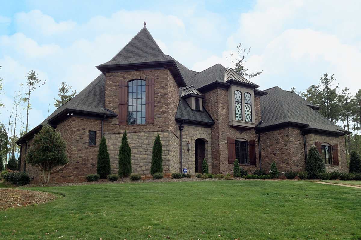 House plan exterior. Two-story brick home with stone accents. Features include multiple gables, arched entryway, and dormers with decorative windows.