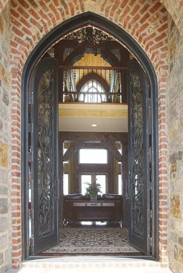 Gothic arched entryway with ornate iron doors opens to a foyer with a balcony and large windows.