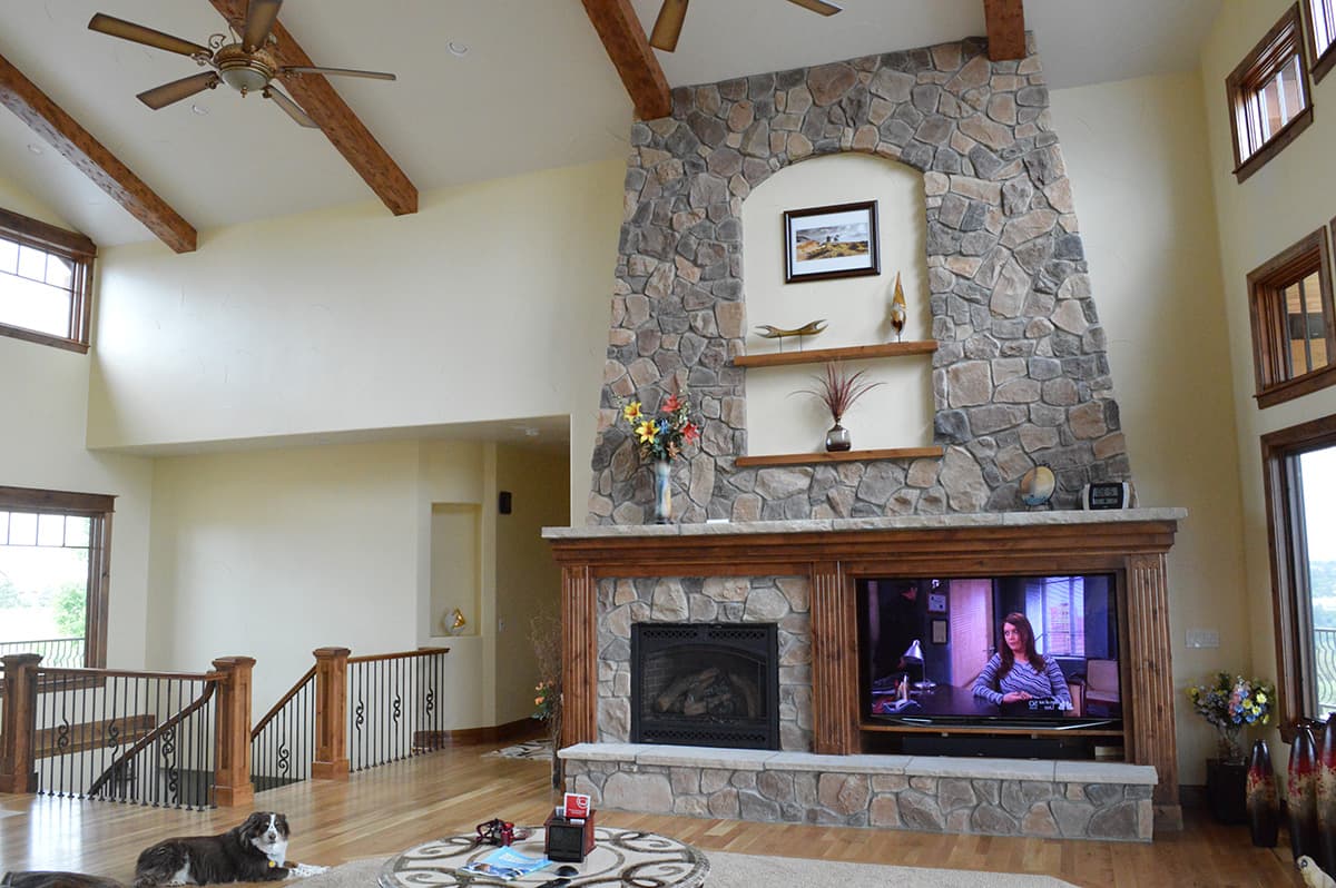 Interior view of a living room with a floor-to-ceiling stone fireplace, vaulted ceiling with wood beams, and a large window.