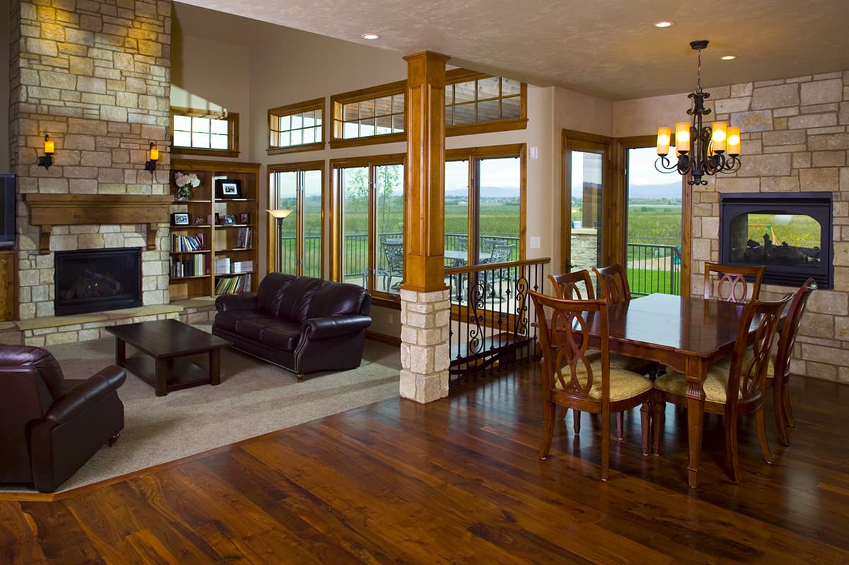 Interior view of a great room with a stone fireplace, dining table, and large windows overlooking a field.
