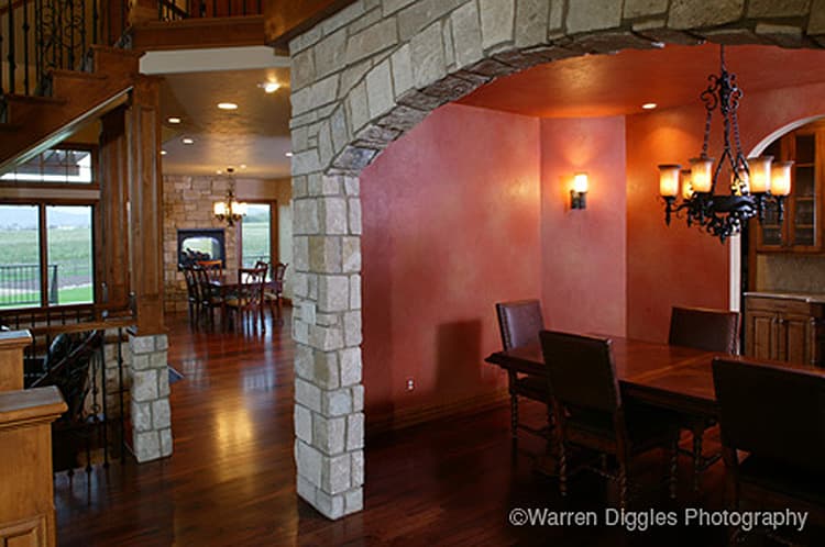 Interior view of a dining room with a stone archway, chandelier, and wood dining set, opening to a living area with a stone fireplace.