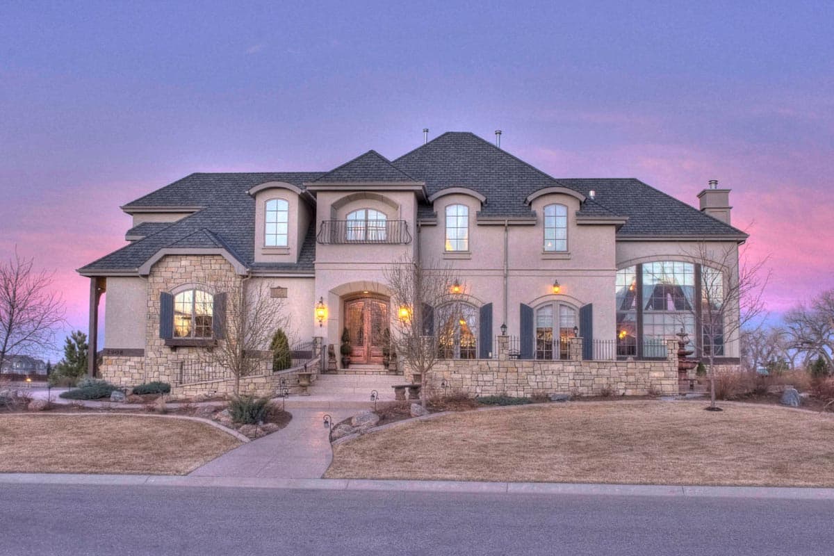 House plan exterior. Two-story home with a stone facade, arched windows with shutters, and a hipped roof. Features include dormers and balcony.