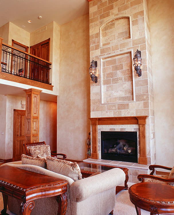 Interior view of a living room with a tall stone fireplace, wood accents, wrought iron balcony railing, and plush seating.