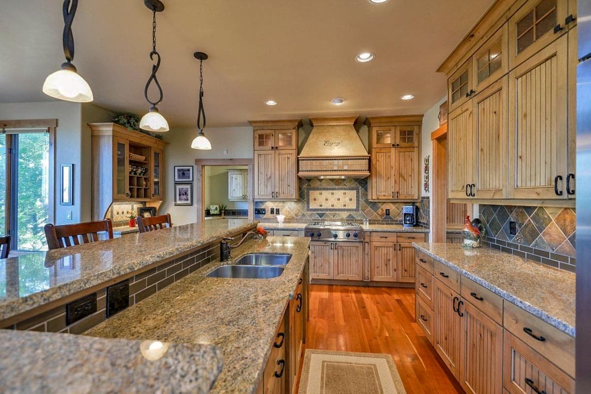 Kitchen interior with a large granite island, stainless steel range, wood cabinets, and pendant lighting.